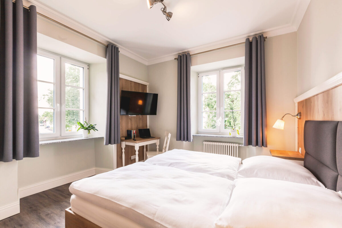 Sonne Nollingen_Apartment_01_2 Helles Schlafzimmer mit einem Doppelbett, weissen Bettdecken und einem Holzkopfteil. Zwei Fenster mit grauen Vorhängen lassen Tageslicht in den Raum. Zwischen den Fenstern befindet sich ein Schreibtisch mit einem Laptop und einem Wandfernseher darüber. Auf der Fensterbank steht eine grüne Pflanze in einem weissen Topf.