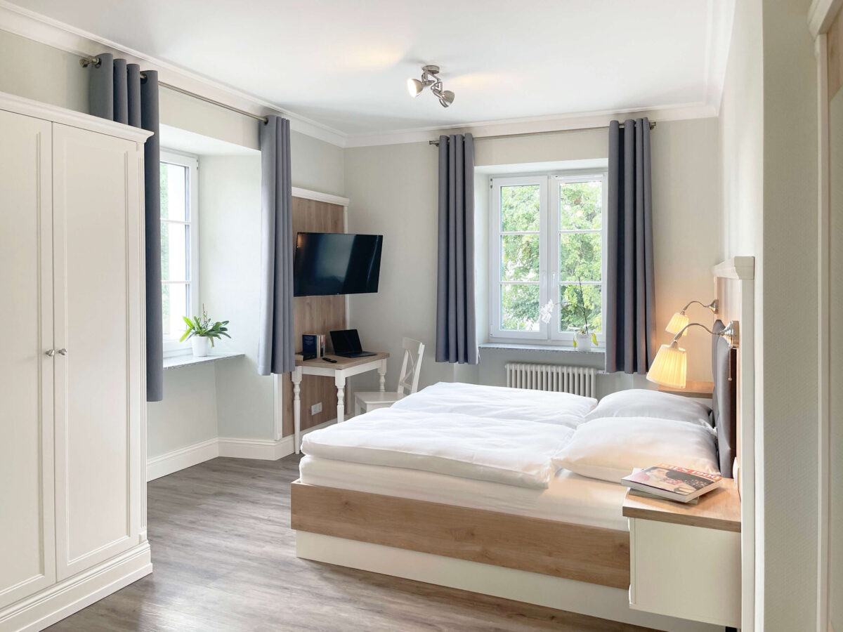 Sonne Nollingen_Apartment_01_sa Helles Schlafzimmer mit einem Doppelbett, weissen Bettdecken und einem Nachttisch mit Leselampe. Ein Fernseher ist an der Wand montiert, und ein kleiner Schreibtisch mit einem Laptop steht daneben. Die Fenster mit grauen Vorhängen lassen Tageslicht in den Raum.