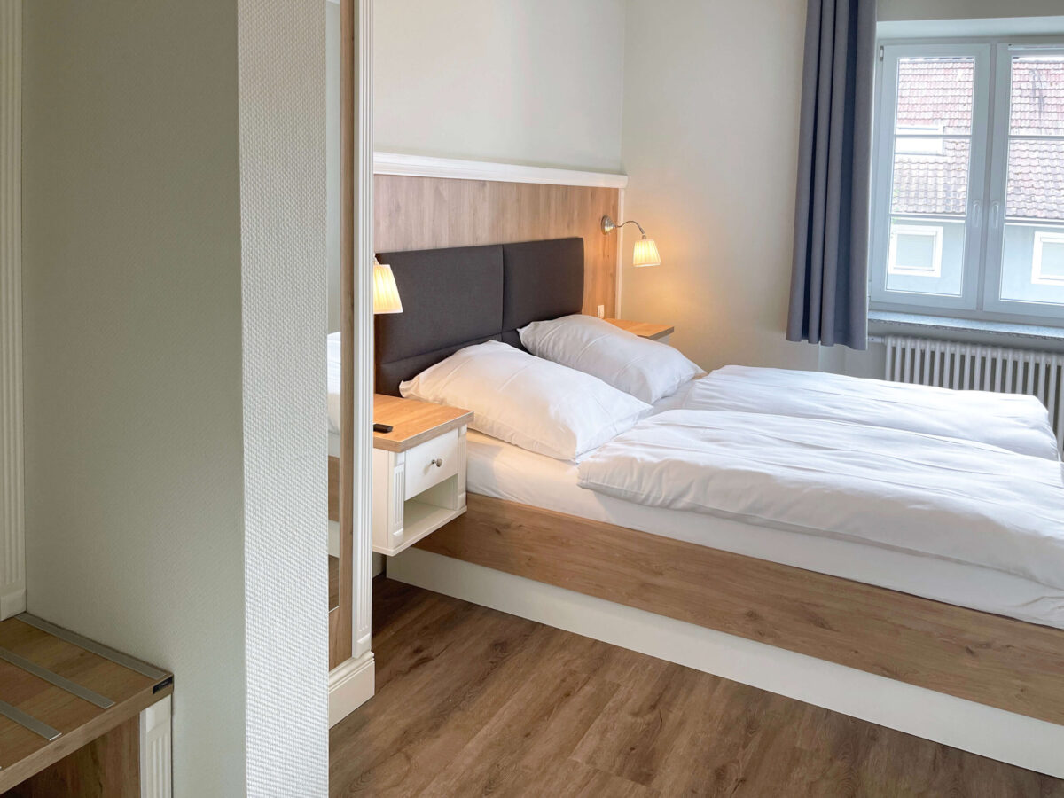Sonne Nollingen_Apartment_02_sa Helles Schlafzimmer mit einem Doppelbett, weissen Bettdecken und einem Nachttisch mit Leselampe. Das Bett hat ein Kopfteil aus Holz und Stoff. Ein Fenster mit grauen Vorhängen lässt Tageslicht in den Raum, auf dem Nachttisch liegt eine Fernbedienung.
