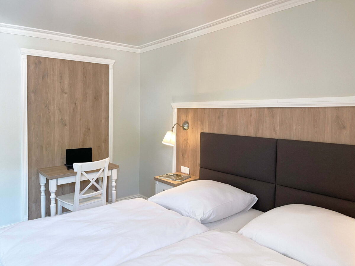 Sonne Nollingen_Apartment_03_sa Schlafzimmer mit einem Doppelbett und einem Kopfteil aus dunkelgrauem Stoff und Holzelementen. Neben dem Bett steht ein Nachttisch mit Leselampe, auf dem Zeitschriften liegen. An der Wand befindet sich ein kleiner Schreibtisch mit einem Laptop und einem weissen Stuhl.