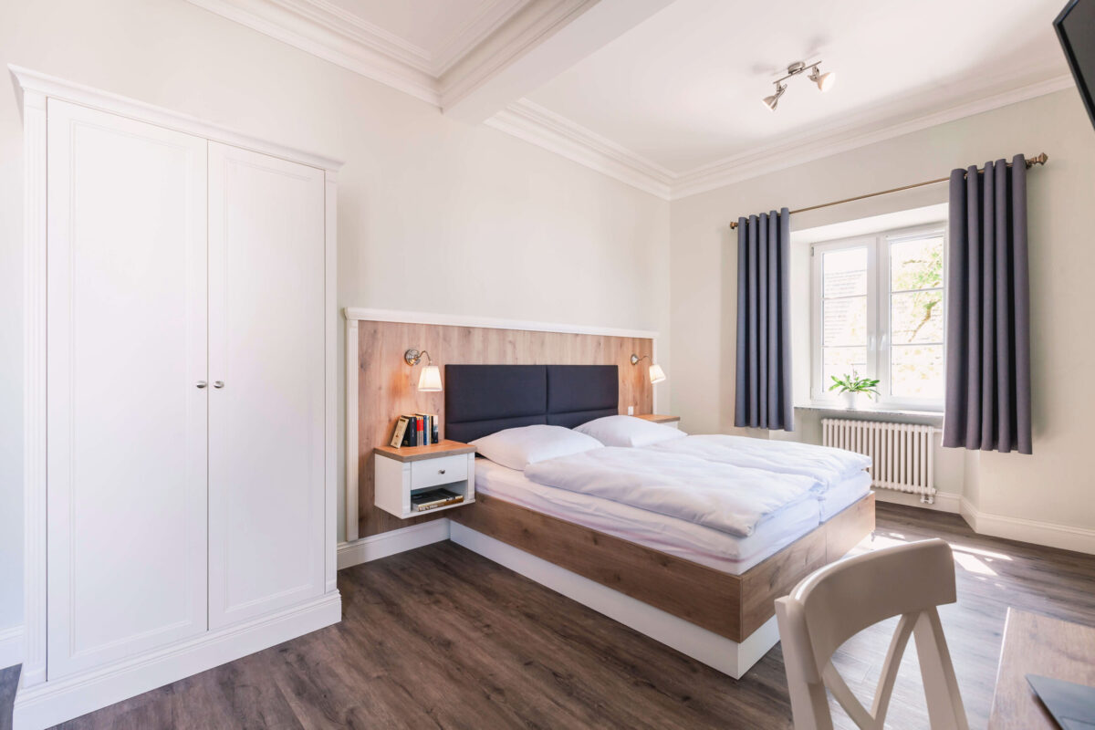 Sonne Nollingen_Apartment_05 Geräumiges Schlafzimmer mit einem Doppelbett, das eine gepolsterte Kopfstütze und integrierte Nachttische mit Büchern und Lampen auf beiden Seiten hat. Der Raum ist in hellen Farben gehalten, mit einem grossen Fenster, das von grauen Vorhängen eingerahmt ist. Rechts neben dem Bett steht ein weisser Kleiderschrank, links ein kleiner Schreibtisch mit Stuhl. Der Boden ist aus dunklem Holzimitat.