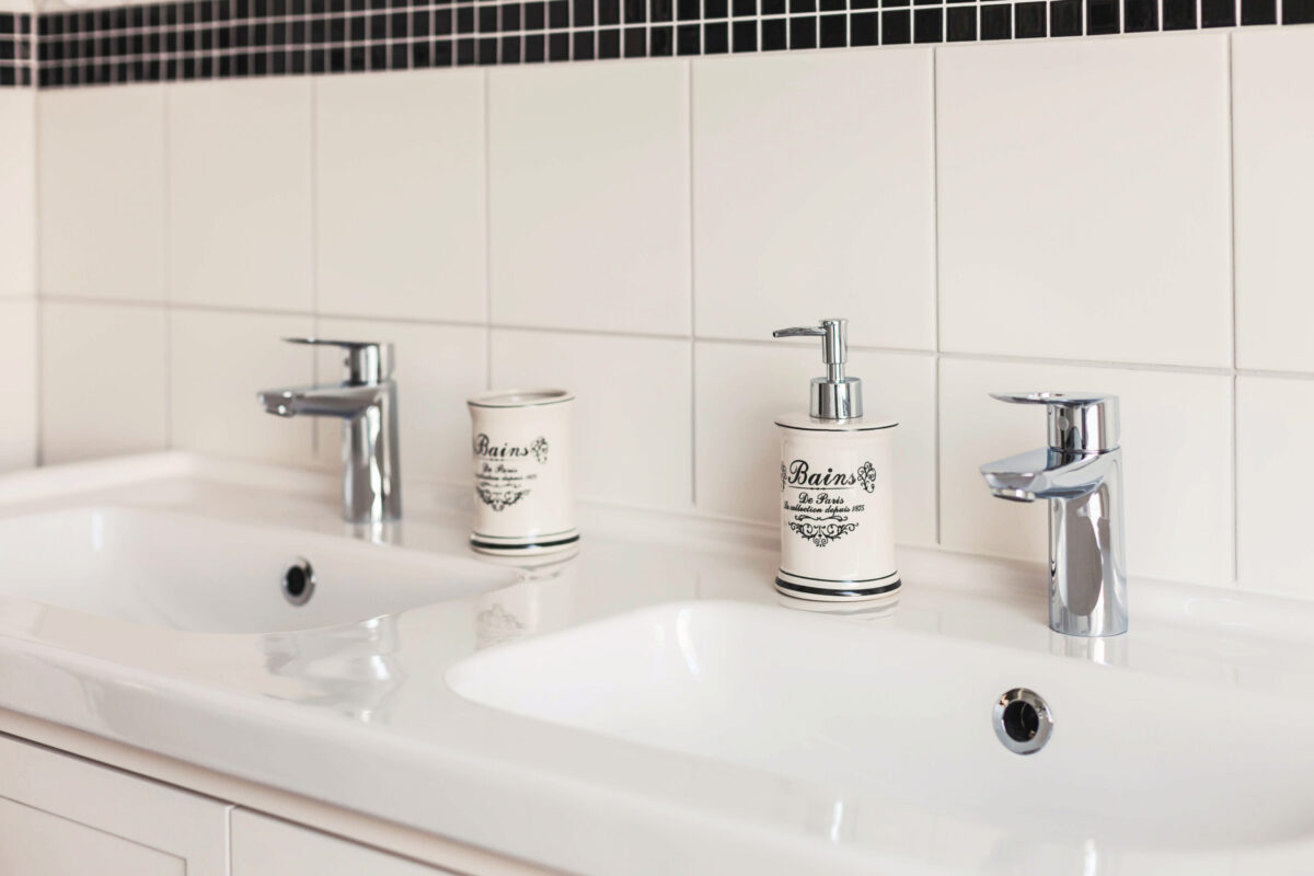 Sonne Nollingen_Badezimmer_03 Detailansicht eines weissen Waschbeckens mit einer modernen Armatur. Links steht ein Zahnputzbecher, rechts ein Seifenspender, beide im Vintage-Stil mit der Aufschrift "Bains de Paris".