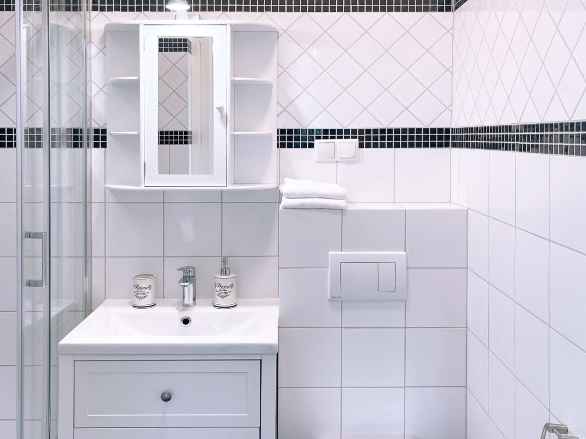 Sonne Nollingen_Badezimmer_04_hoch Kleines, helles Badezimmer mit weissen Fliesen und schwarzen Mosaikakzenten. Ausgestattet mit einer Duschkabine, einem Waschbecken mit Spiegel und Unterschrank sowie einer Toilette. Handtücher und Hygieneartikel sind ordentlich aufgestellt.