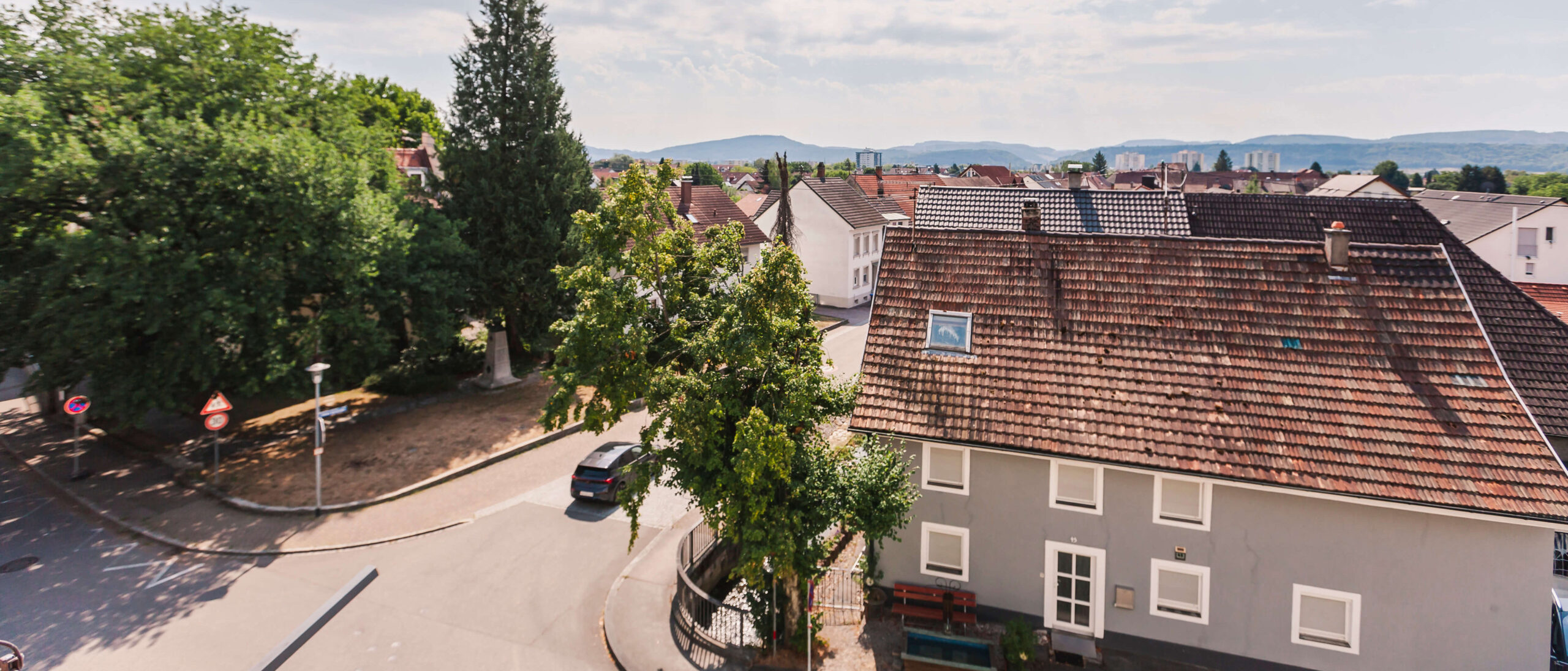 Aussicht auf eine ruhige Wohngegend mit mehreren Häusern, von denen eines ein graues Haus mit roten Dachziegeln ist. Im Vordergrund sind grosse Bäume und eine Strassenkreuzung zu sehen. Im Hintergrund erstreckt sich eine hügelige Landschaft unter einem leicht bewölkten Himmel.