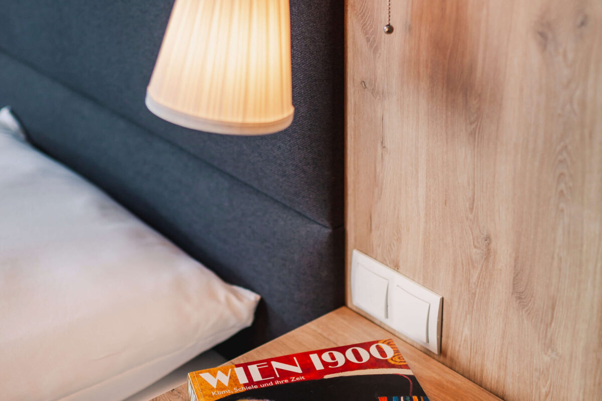 Sonne Nollingen_Impressionen_04_hoch Nachttisch mit einer eingeschalteten Leselampe an der Wand. Auf dem Tisch liegt ein Buch mit dem Titel "Wien 1900" und einer Abbildung von Gustav Klimts Gemälde "Die Frau in Gold".