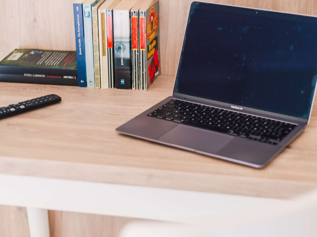 Sonne Nollingen_Impressionen_05_hoch Holzschreibtisch mit einem aufgeklappten Laptop und einer Fernbedienung. Neben dem Laptop steht eine kleine Sammlung von Büchern, darunter ein Buch von Stieg Larsson.