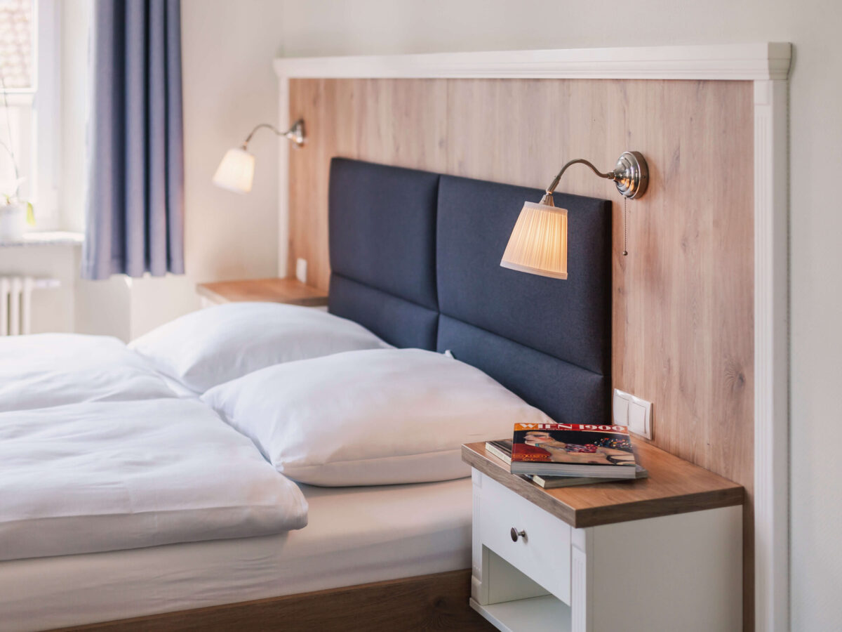 Sonne Nollingen_Impressionen_07 Ein ordentlich gemachtes Bett mit gepolstertem Kopfteil und zwei Wandlampen. Auf einem kleinen Nachttisch liegen zwei Zeitschriften. Im Hintergrund ist ein Fenster mit blauen Vorhängen zu sehen.