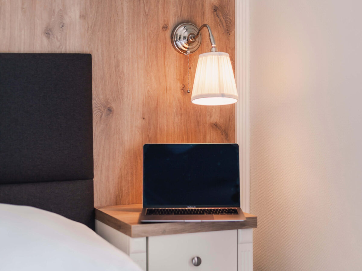 Sonne Nollingen_Impressionen_08_hoch Kleiner Nachttisch neben einem Bett mit einem aufgeklappten Laptop darauf. Darüber hängt eine eingeschaltete Leselampe an einer Holzwand.