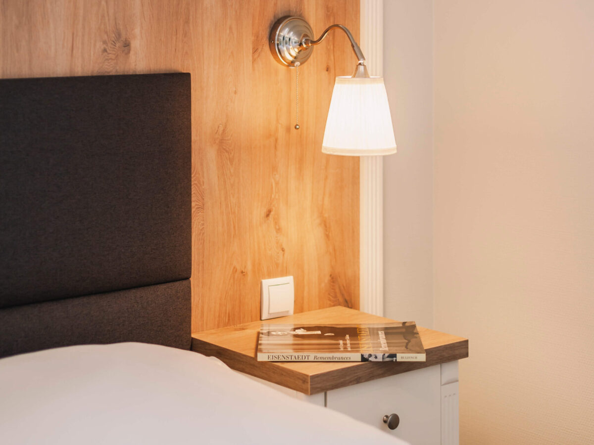 Sonne Nollingen_Impressionen_10_hoch Nachttisch mit einer eingeschalteten Wandlampe über einem Bett. Auf dem Tisch liegt ein Buch mit dem Titel "Eisenstaedt Remembrances". Die Wand hinter dem Bett ist mit Holzelementen verkleidet.