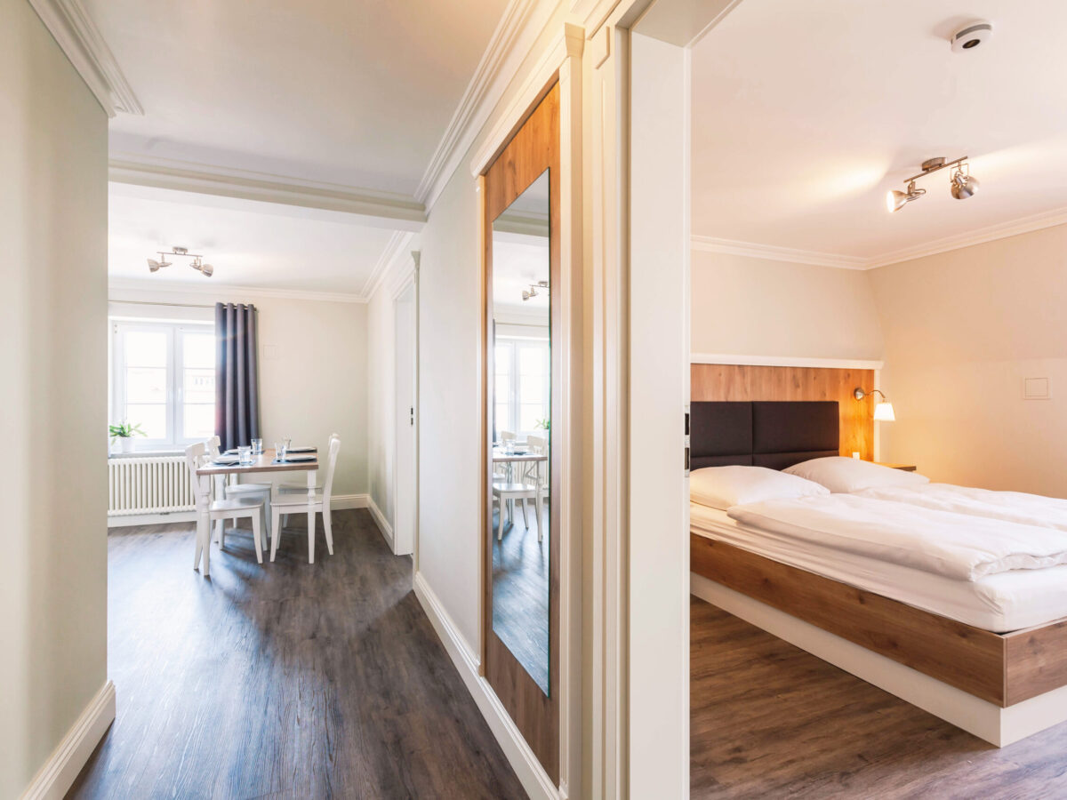 Sonne Nollingen_Impressionen_13 Helles Apartment mit einem offenen Flur, der zu einem Schlafzimmer und einem Essbereich führt. Das Schlafzimmer hat ein Doppelbett mit Holzelementen und Leselampen. Der Essbereich ist mit einem Tisch und weissen Stühlen vor einem Fenster mit grauen Vorhängen ausgestattet. Der Boden besteht aus dunklem Holzlaminat, und im Flur hängt ein grosser Wandspiegel.