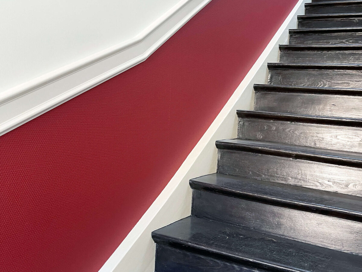 Sonne Nollingen_Impressionen_14_hoch Dunkle Holztreppe mit einer rot tapezierten Wand und weissen Zierleisten. Die Wand entlang der Treppe ist im oberen Bereich weiss gestrichen.