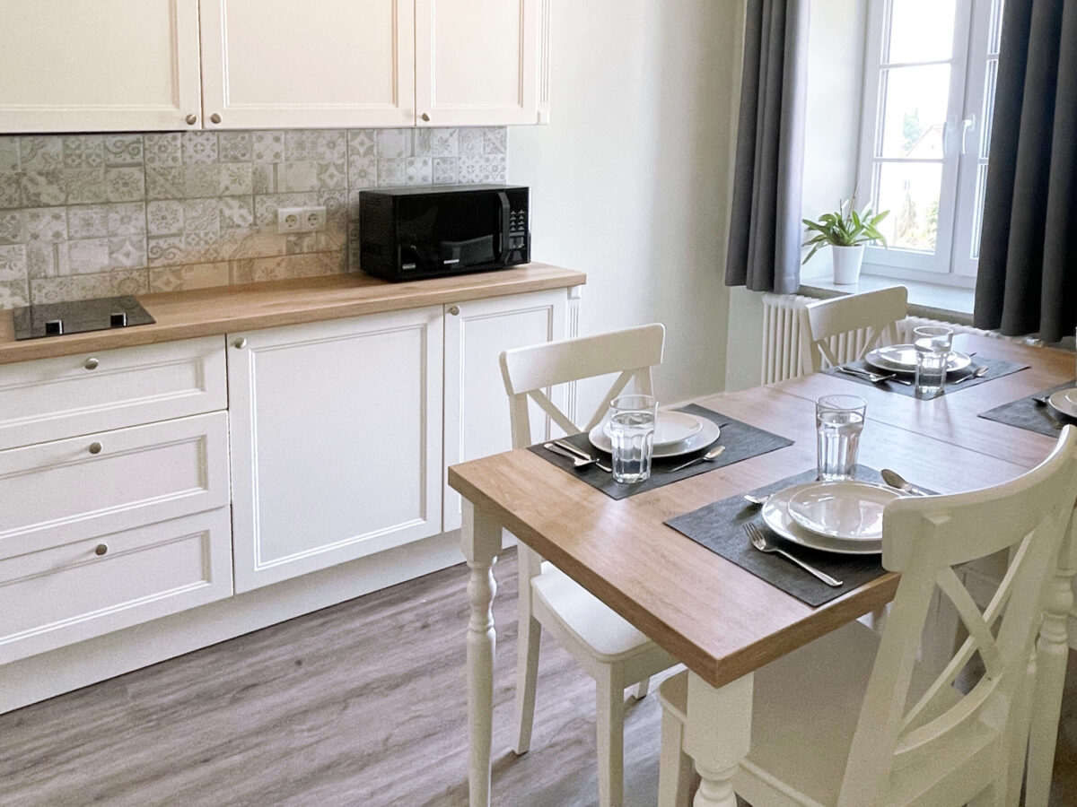 Sonne Nollingen_Kueche_Family_02_sa Helle Küche mit weissen Schränken, Holzarbeitsplatten und gemusterten Fliesen. Auf der Arbeitsplatte steht eine Mikrowelle, und das Kochfeld ist in die Arbeitsfläche integriert. Vor einem Fenster mit grauen Vorhängen steht ein gedeckter Esstisch mit weissen Stühlen, Tellern, Gläsern und Besteck.