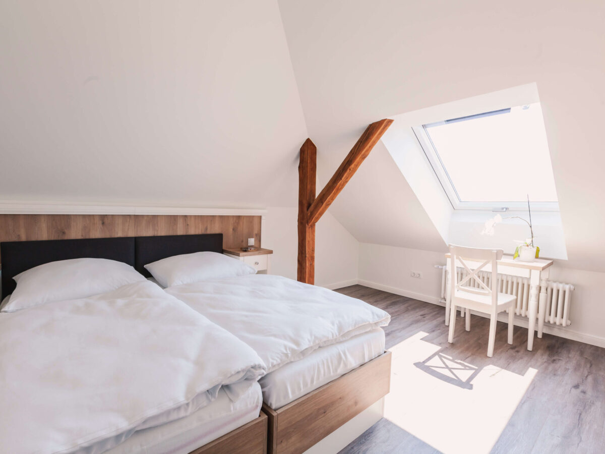 Sonne Nollingen_Penthouse_02 Helles Schlafzimmer im Dachgeschoss mit einem Doppelbett, weissen Bettdecken und Holzelementen. Ein Holzbalken teilt den Raum, und ein Schreibtisch mit Stuhl steht vor einem Dachfenster, durch das viel Tageslicht einfällt. Auf dem Schreibtisch steht eine weisse Orchidee.