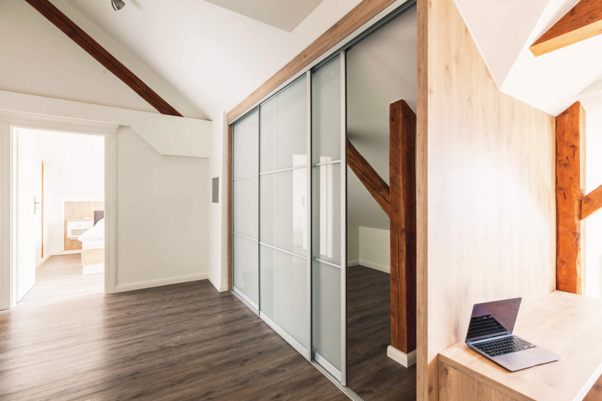 Sonne Nollingen_Penthouse_03 Geräumiger Flur mit Dachschrägen, sichtbaren Holzbalken und einem grossen Kleiderschrank mit Schiebetüren aus Milchglas. Auf der rechten Seite steht ein Holzschreibtisch mit einem offenen Laptop. Im Hintergrund ist ein Schlafzimmer zu sehen.