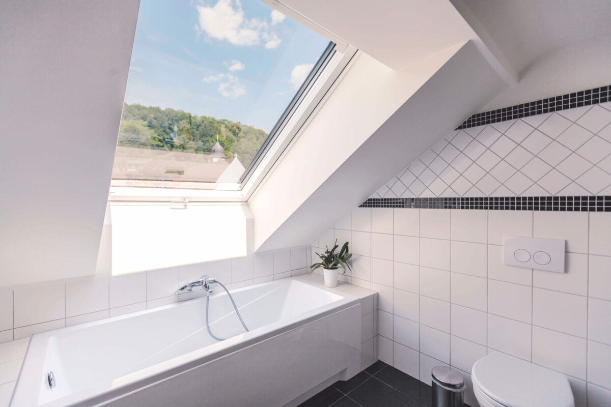 Sonne Nollingen_Penthouse_Bad_01 Modernes Badezimmer mit Badewanne unter einem Dachfenster, das einen Blick ins Grüne bietet. Die Wände sind weiss gefliest mit einem dekorativen Mosaikband, daneben befindet sich eine Toilette und eine kleine Pflanze auf dem Wannenrand.