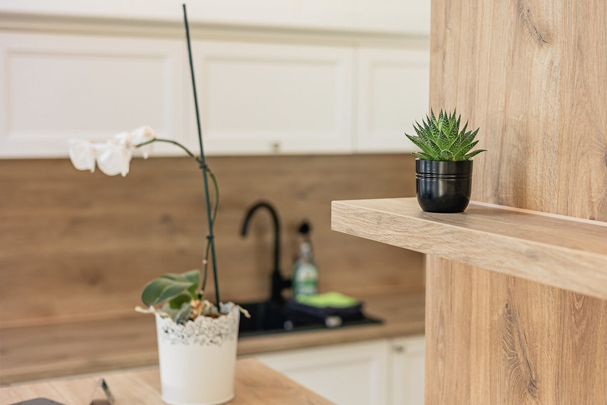 sonne-nollingen-apartmenthotel-penthouse-wohnung-impressionen-02 Detailansicht einer modernen Küche mit Holzakzenten. Im Vordergrund steht eine Topfpflanze auf einem Regal, daneben eine Orchidee in einem weissen Topf auf der Arbeitsfläche. Im Hintergrund ist eine weisse Küchenzeile mit Holzrückwand und ein schwarzer Wasserhahn zu sehen.