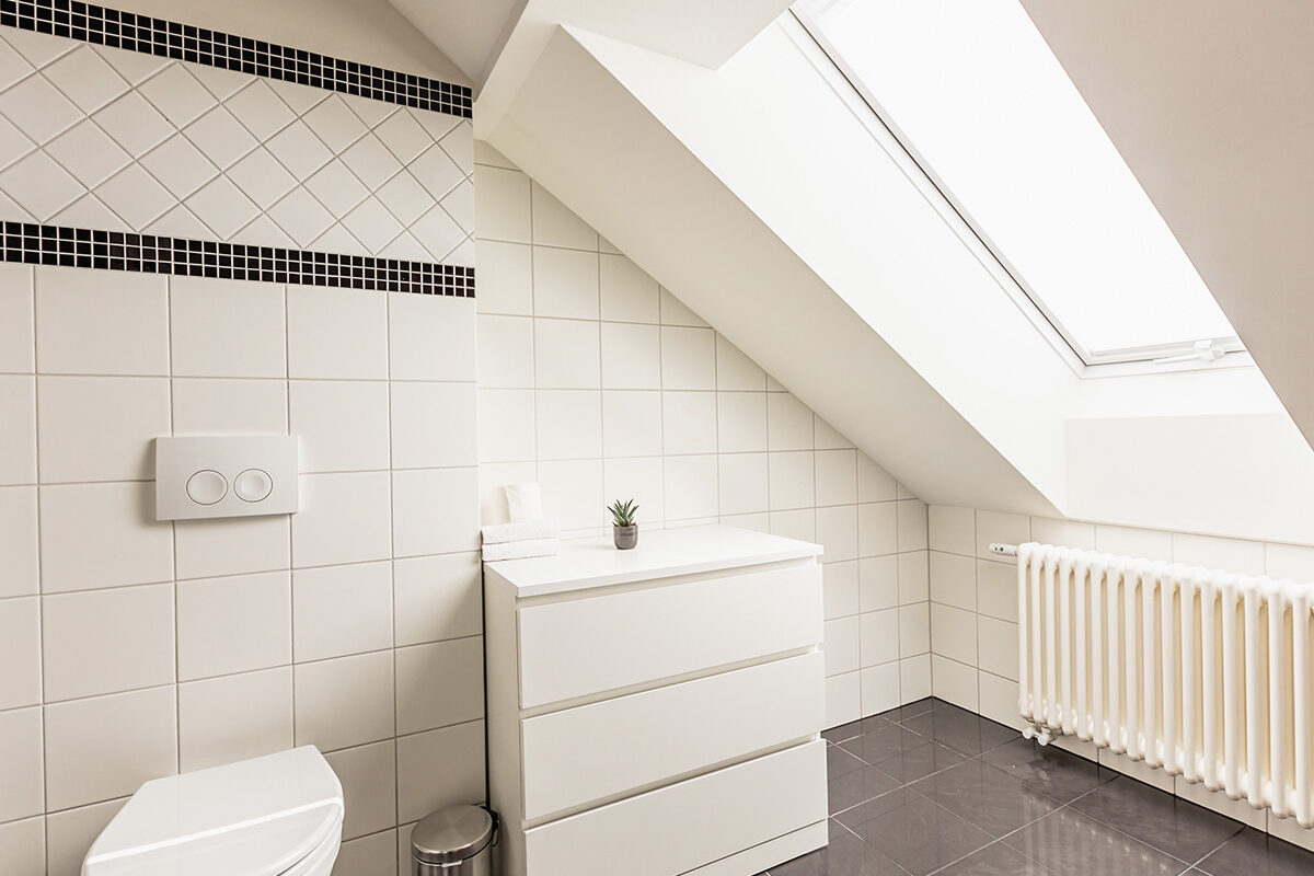 sonne-nollingen-apartmenthotel-penthouse-wohnung-impressionen-03 Helles Badezimmer mit Dachschräge und Dachfenster. Weisse Fliesen mit schwarzem Mosaikband, ein weisses WC und eine schlichte Kommode mit Handtüchern und einer kleinen Topfpflanze. Der Boden ist dunkel gefliest, ein weisser Heizkörper befindet sich unter dem Fenster.