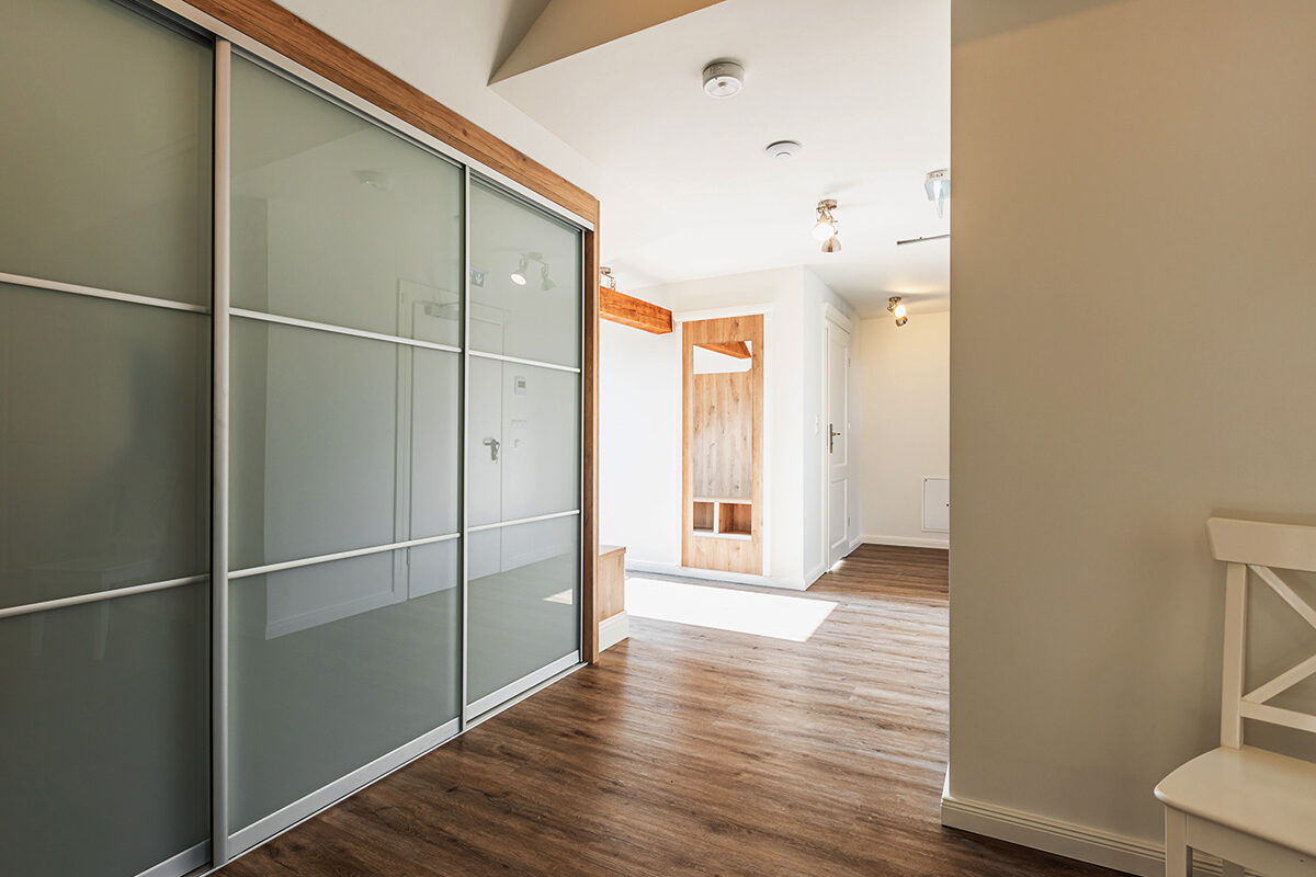 sonne-nollingen-apartmenthotel-penthouse-wohnung-impressionen-04 Moderner Flur mit grossen Einbauschränken mit matten Glastüren und Holzrahmen. Der Boden ist mit dunklem Holzlaminat ausgelegt, eine weisse Stuhl steht am Rand. Im Hintergrund ein heller Eingangsbereich mit Holzdetails und mehreren Türen.