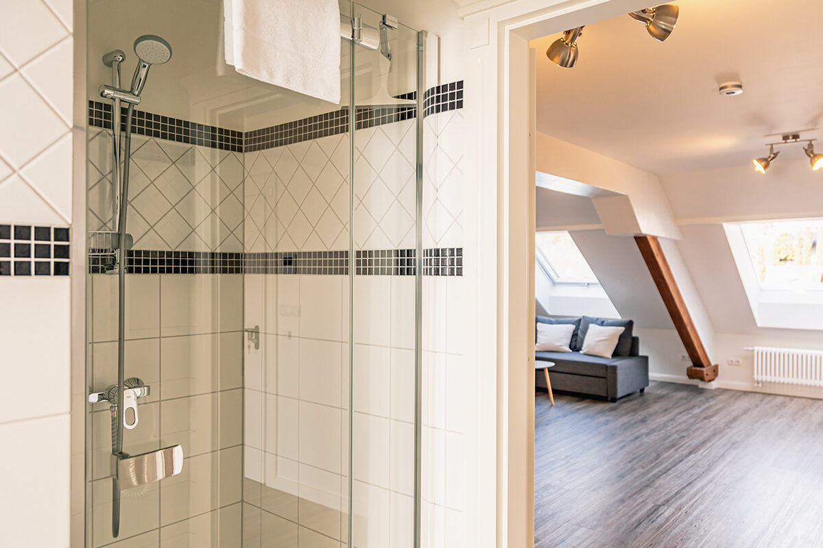sonne-nollingen-apartmenthotel-penthouse-wohnung-impressionen-05 Blick in ein Badezimmer mit einer verglasten Duschkabine. Die Wände sind mit weißen Fliesen und einer schwarzen Mosaik-Bordüre gestaltet. Im Hintergrund ist ein Raum mit Dachschrägen, Holzbalken und einem grauen Sofa zu sehen, beleuchtet durch Deckenstrahler und ein großes Dachfenster.