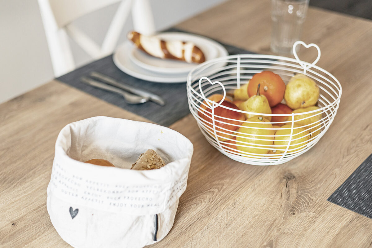 sonne-nollingen-apartmenthotel-penthouse-wohnung-impressionen-18 Detailaufnahme eines gedeckten Esstisches mit einem Brotkorb aus Stoff, gefüllt mit Brötchen, und einem weissen Drahtkorb mit Äpfeln und Birnen. Im Hintergrund ist ein Teller mit einer Brezel sowie ein Glas zu sehen.